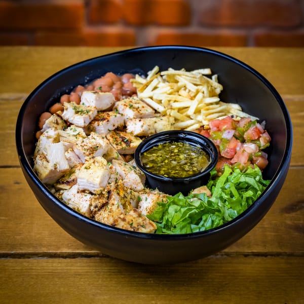 A black bowl filled with grilled chicken, beans, french fries, salsa, lettuce, and a side of green sauce.