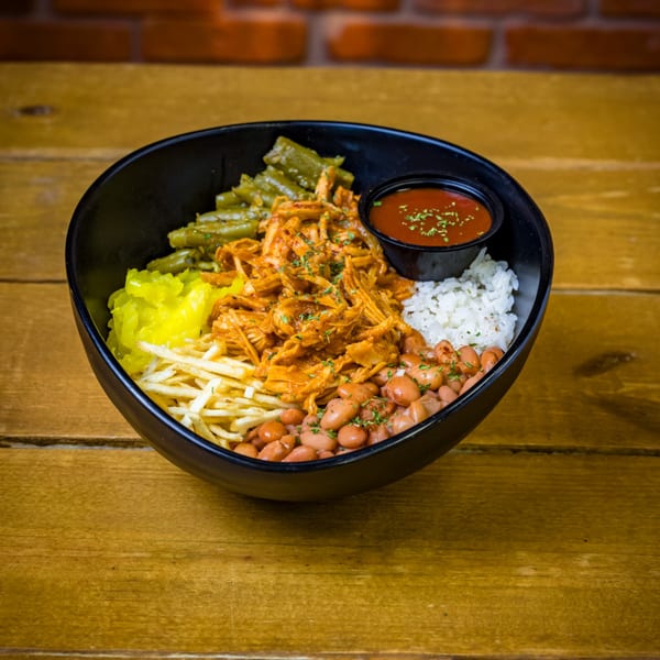A black bowl filled with shredded chicken, green beans, rice, beans, fries, and a side of sauce.