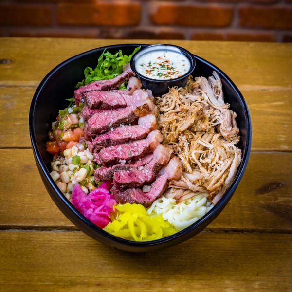 A black bowl filled with sliced steak, pulled pork, corn salsa, pickled onions, and a side of sauce.